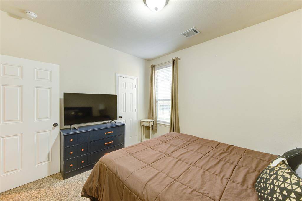 This cozy bedroom features a neutral color palette, ample natural light from a window with curtains, and includes a TV on a dresser. It has a soft carpeted floor and a comfortable bed, making it an inviting space.