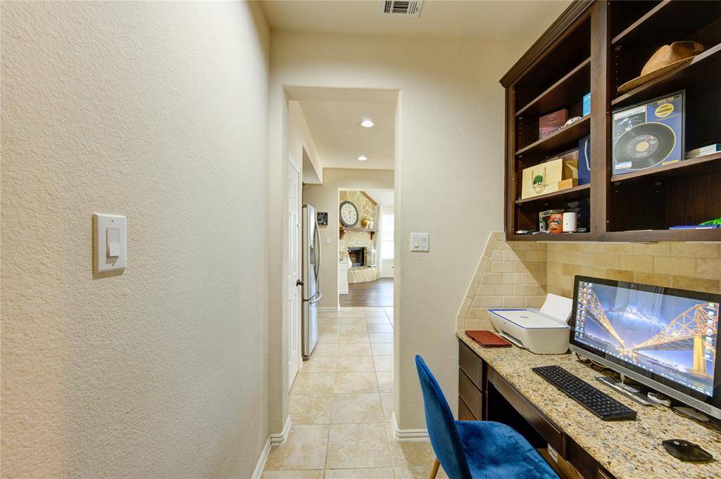 This photo showcases a cozy home office nook with built-in shelves and a granite desk. It opens into a hallway leading to a bright kitchen with stainless steel appliances and a view of a living area with a fireplace. The space features a mix of tile and wood flooring, creating a warm and functional layout.