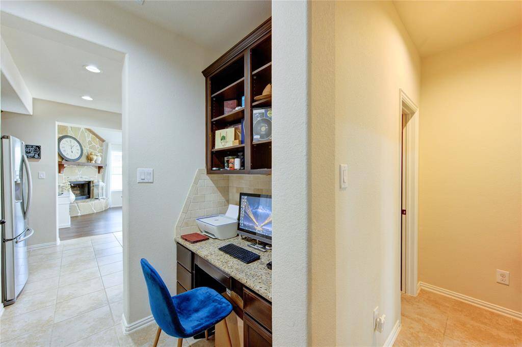 This photo showcases a cozy workspace nook with built-in shelving and a desk, adjacent to a hallway and kitchen. The kitchen features modern appliances and tile flooring, while the view extends to a living area with a stone fireplace and wooden floors.