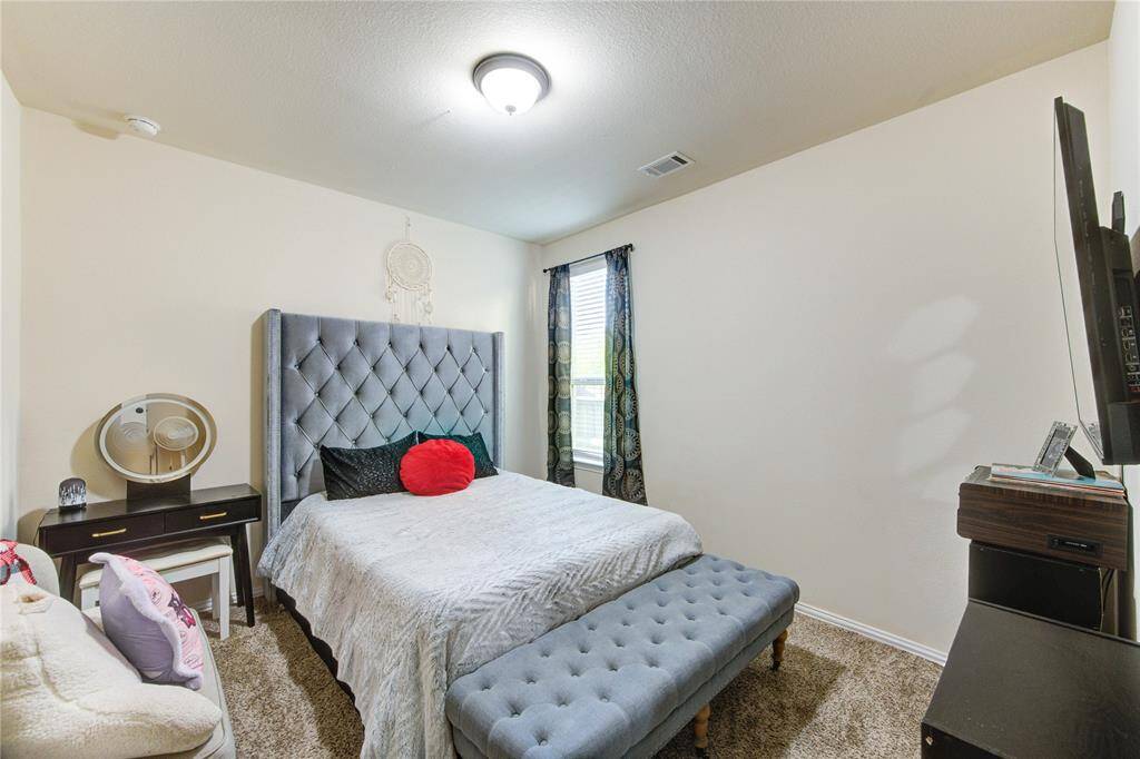 Cozy bedroom with a plush, tufted headboard and matching bench. Features a desk with a round mirror, carpeted floor, and a wall-mounted TV. Natural light filters through a window with patterned curtains.