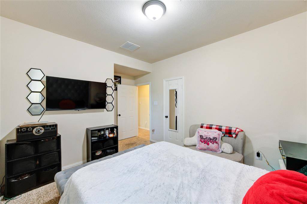 Cozy bedroom with neutral walls, a ceiling light, and carpeted floor. Features include a wall-mounted TV, a small sofa with decorative pillows, and a mirror on the door. The space is well-organized with additional storage under the TV.