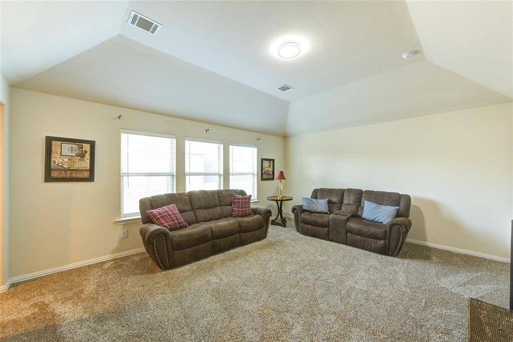 This spacious living room features plush carpeting, neutral walls, and ample natural light from three large windows. It includes two comfortable brown sofas with accent pillows and a small side table with a lamp, creating a cozy and inviting atmosphere.