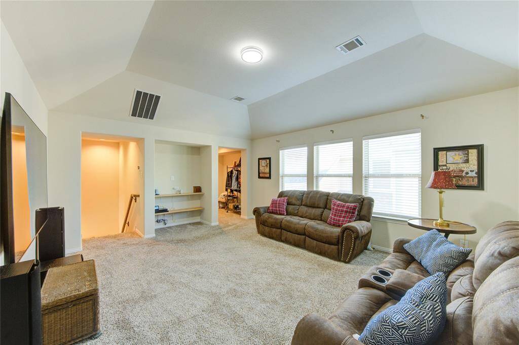 This spacious living room features plush carpeting, vaulted ceilings, and ample natural light from multiple windows. It includes comfortable seating and a cozy entertainment area, with built-in shelving and a small adjacent nook.