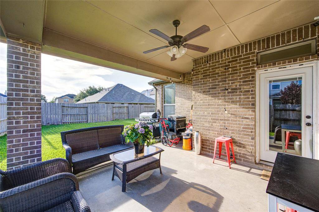 This photo showcases a covered patio with brick walls, featuring a ceiling fan, comfortable seating, a coffee table, and a view of a fenced backyard, perfect for outdoor relaxation and entertaining.