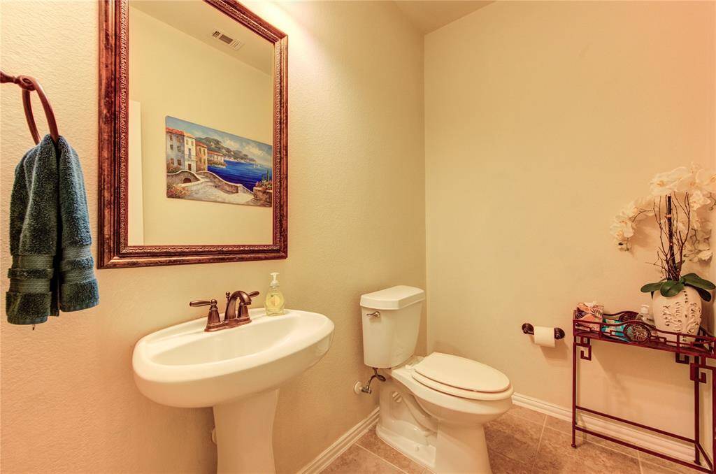This bathroom features a classic design with a pedestal sink, wall-mounted mirror, and neutral tones. It includes a toilet, towel rack, decorative artwork, and a small shelf with floral arrangements, creating an inviting and functional space.