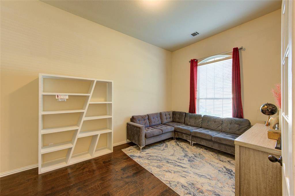 Cozy and well-lit room featuring dark hardwood floors, a modern L-shaped sofa, and a unique angled bookshelf. A large window with red curtains allows natural light to fill the space, highlighting the elegant patterned rug and sleek side table. Perfect for a comfortable living area or home office.