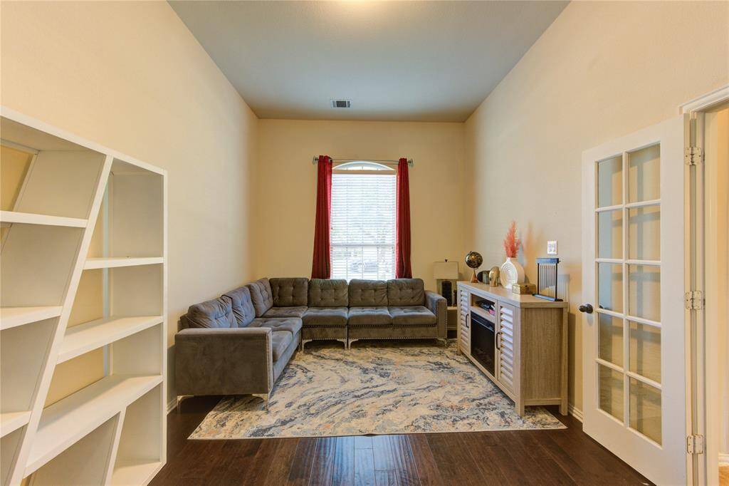 This cozy living room features high ceilings, a large window with red curtains, and a sectional sofa. It includes a decorative rug, built-in shelving, and a stylish console with a fireplace. French doors add elegance, and the warm color palette creates an inviting atmosphere.