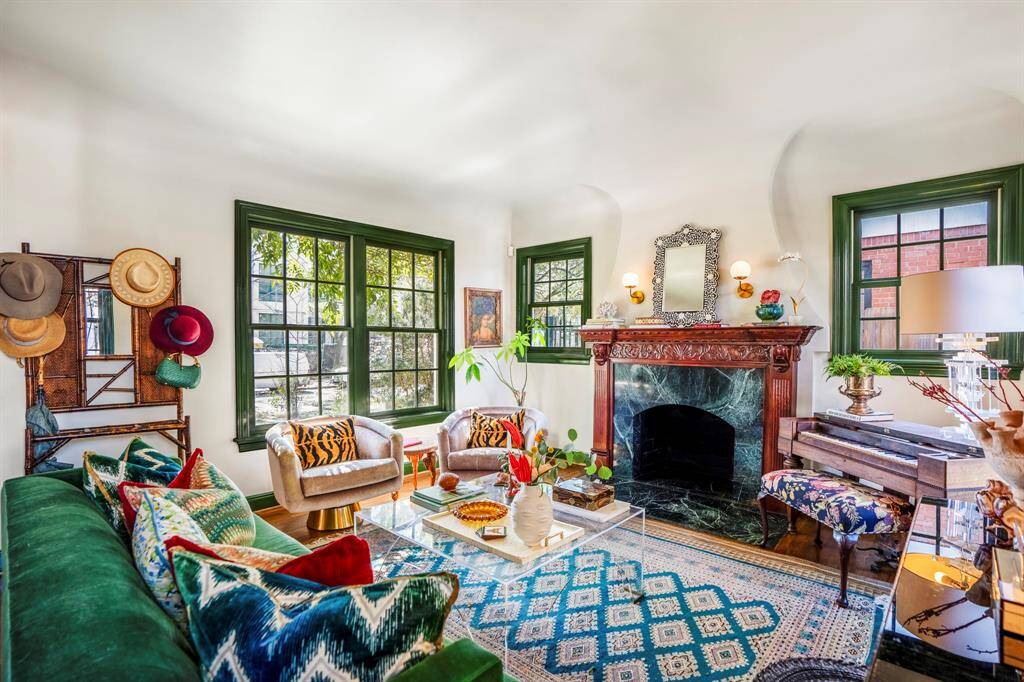 Sunlight pours through green-painted windows, casting a warm glow over the original marble fireplace. A space designed for conversation, laughter, and quiet moments by the fire.