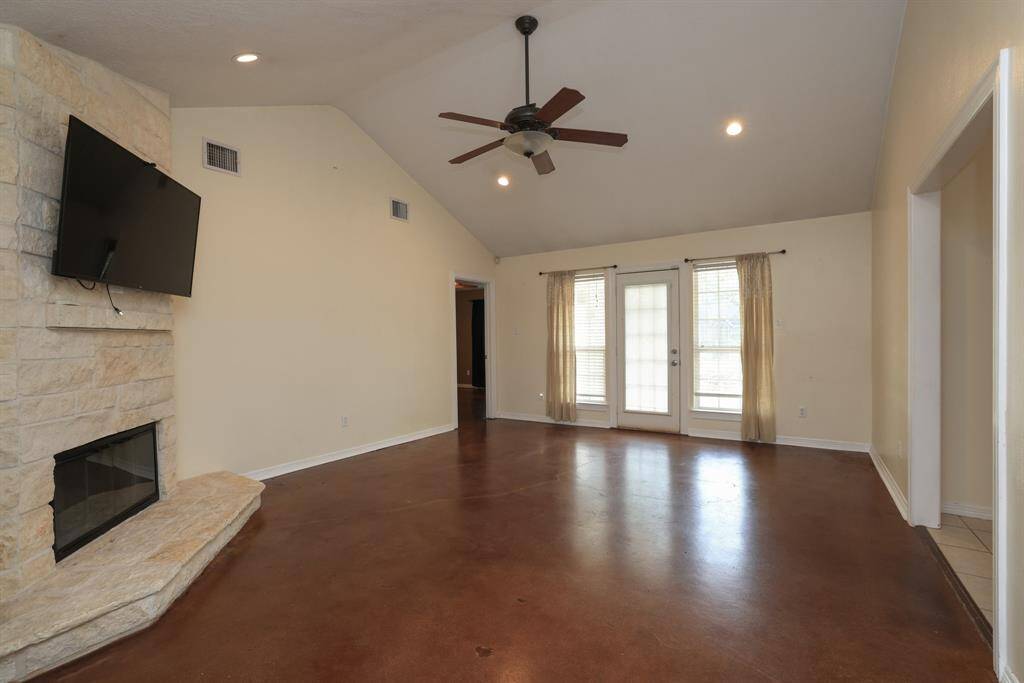 This spacious living room features a high ceiling with a central fan, a stone fireplace,  and large windows with double doors leading outside, providing ample natural light.
