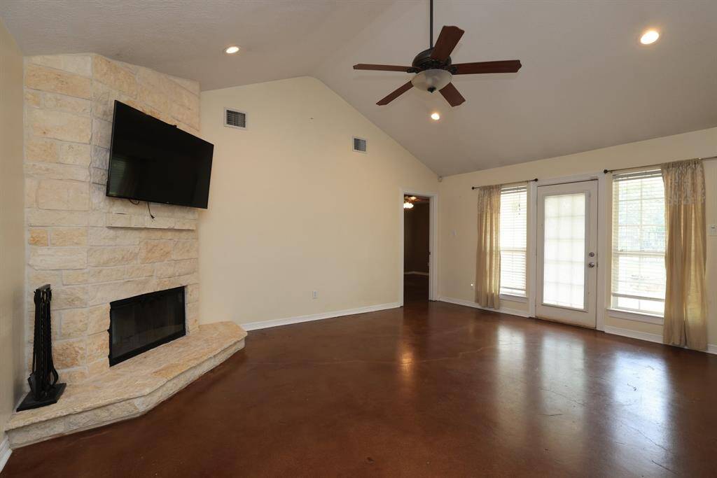 This spacious living room features a vaulted ceiling, ceiling fan, and a stone fireplace. The polished concrete floor and neutral walls create a modern, open feel, while the entryway provides natural light.