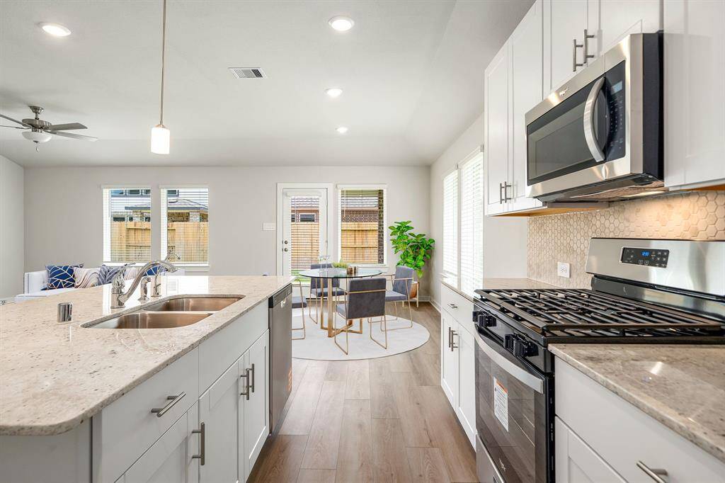 This stunning kitchen boasts gleaming granite countertops, a large center island with double sink, and contemporary white cabinetry complemented by sleek stainless steel appliances. The stylish hexagonal tile backsplash adds a touch of elegance, while expansive windows and a glass-paneled door fill the space with natural light, creating a warm and inviting atmosphere.