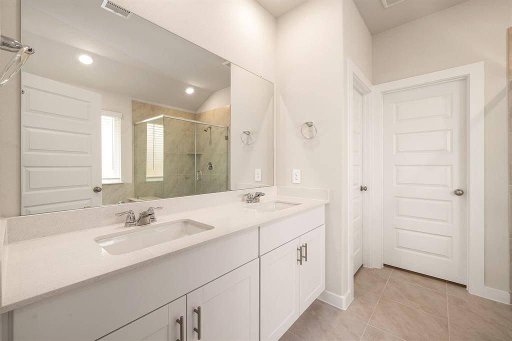 This elegant bathroom features a modern double-sink vanity with sleek quartz countertops, contemporary fixtures, and ample storage. The spacious walk-in shower is enclosed in glass and lined with neutral tile, complementing the soft-toned walls and stylish flooring. Clean lines and natural light enhance the bright, airy atmosphere—perfect for a relaxing retreat.