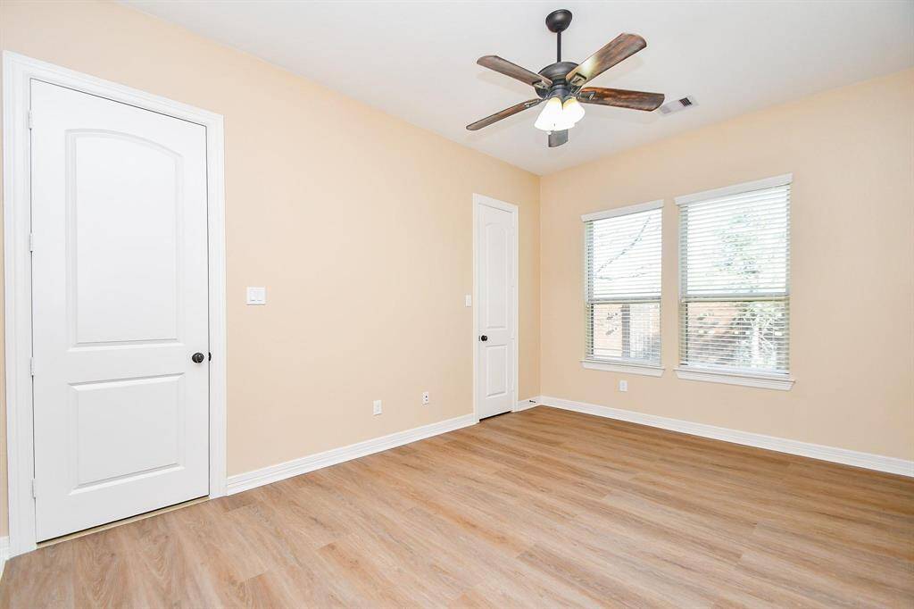 This room features neutral walls, wood-like flooring, and ample natural light from two large windows. It includes a ceiling fan and two white doors, offering a clean and versatile space.