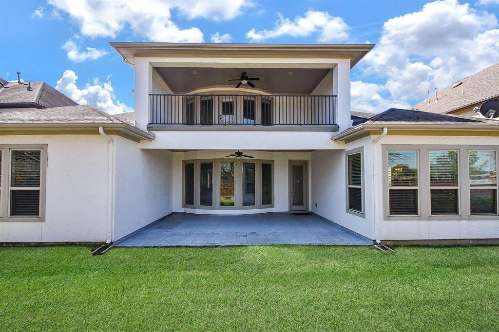 This photo showcases the back of a modern home with a spacious covered patio and well-maintained lawn. The upper level features a balcony with a ceiling fan, overlooking the yard, perfect for outdoor relaxation. Large windows provide ample natural light to the interior.