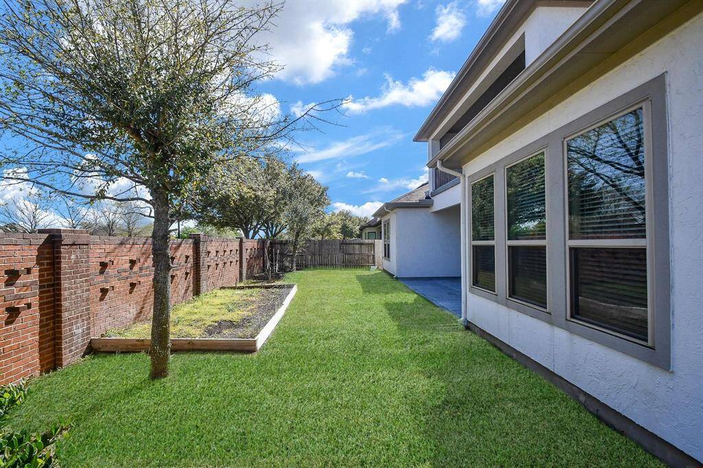 The photo shows a well-maintained backyard featuring a lush green lawn, a brick privacy wall, and a few trees for shade. The exterior of the home has multiple large windows, allowing plenty of natural light. Ideal for outdoor activities and relaxation.
