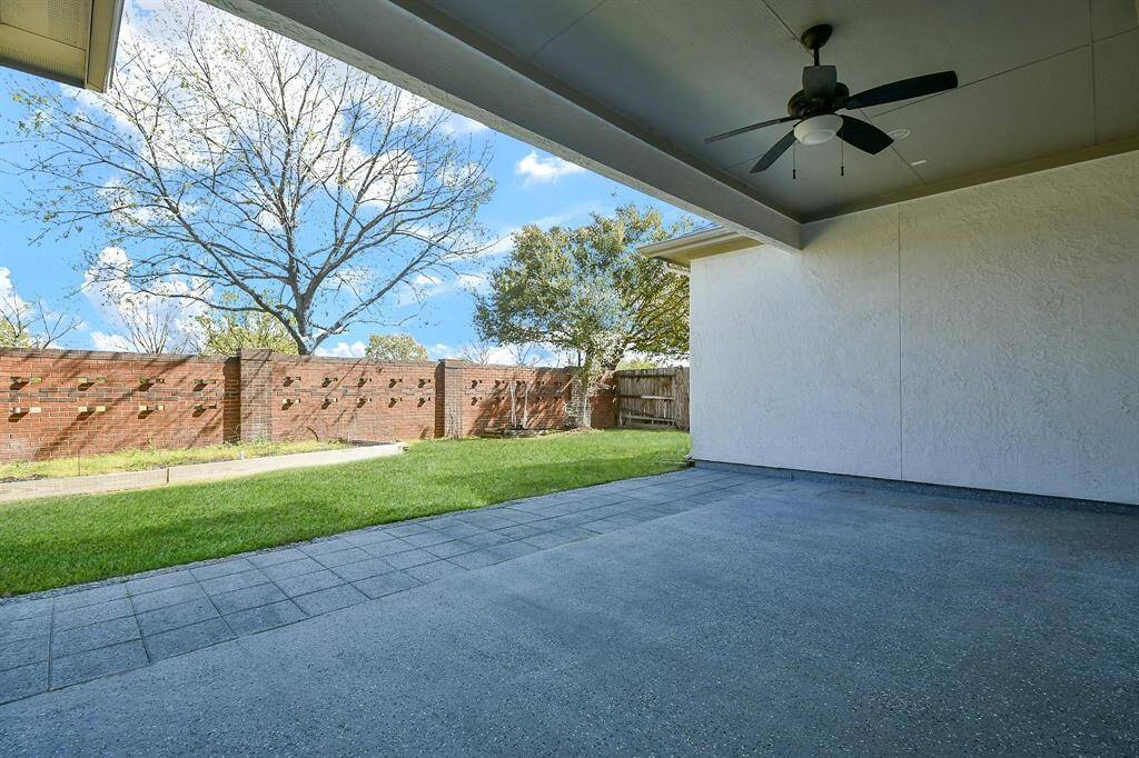 This photo showcases a covered patio with a ceiling fan, overlooking a spacious backyard with a brick fence and mature trees, perfect for outdoor relaxation or entertaining.