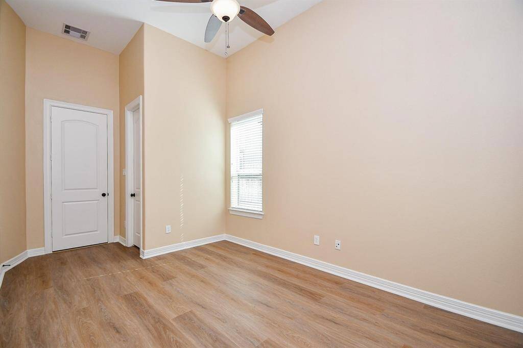 This room features neutral-toned walls, wood flooring, and a ceiling fan. It has a bright window and a white door, offering a clean and airy feel, ideal for a bedroom or office space.