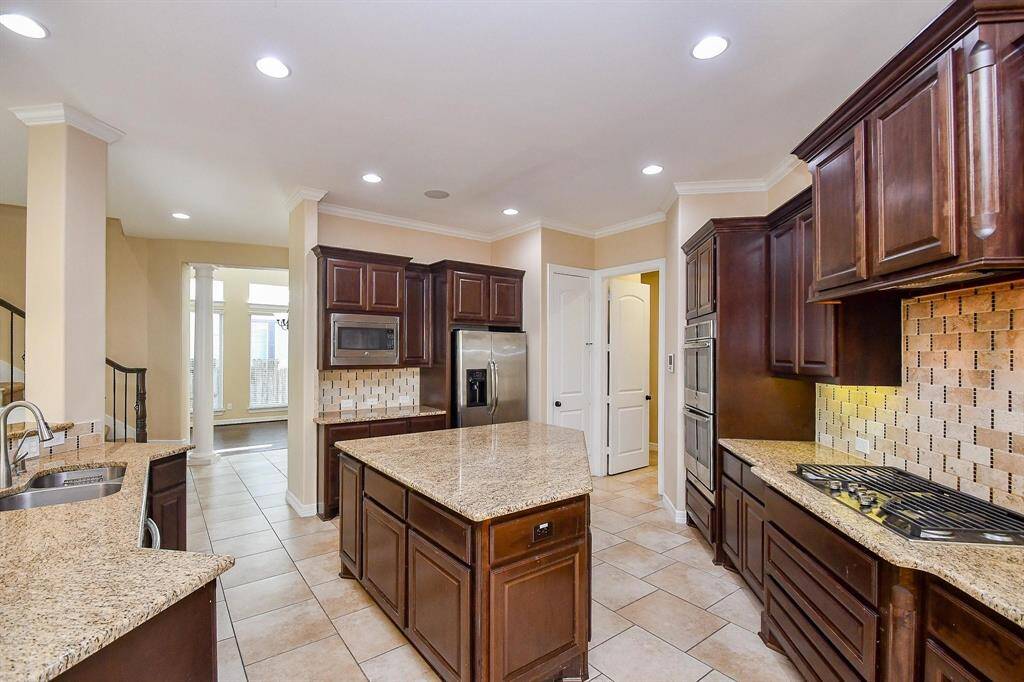This kitchen features rich, dark wood cabinetry with granite countertops and a stylish tile backsplash. It includes stainless steel appliances, a built-in microwave, and a spacious island, perfect for cooking and entertaining.