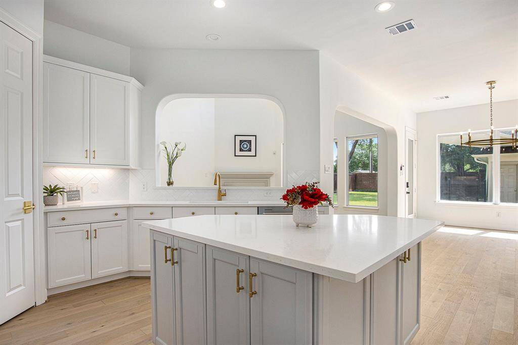 Bright and spacious kitchen with modern white cabinetry, a large central island, and elegant gold hardware. The open layout flows into a sunlit dining area with large windows offering views of the backyard. Perfect for entertaining and family gatherings.