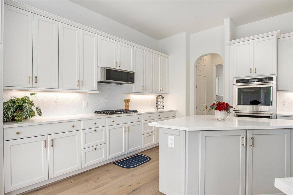 This modern kitchen features sleek white cabinetry, a spacious island, stainless steel appliances, and under-cabinet lighting. The light wood flooring and neutral color palette create an inviting and airy atmosphere.
