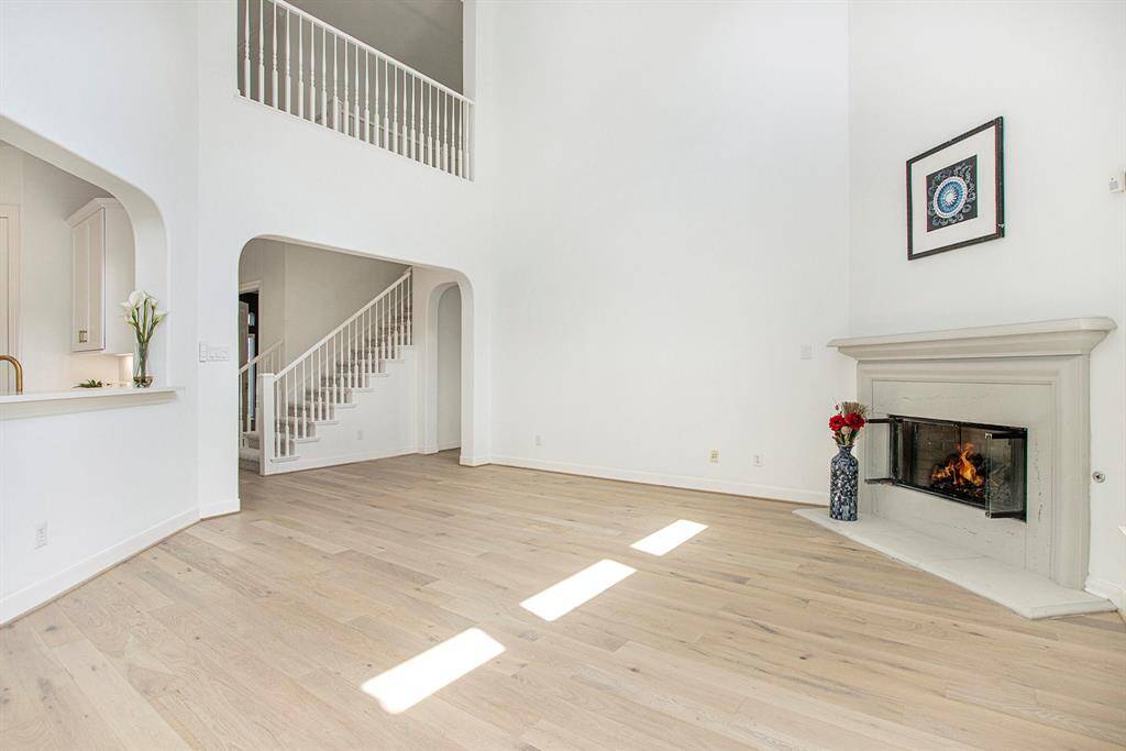 This spacious living area features high ceilings, a cozy gas log fireplace, and light wood flooring. The open layout includes a view of the staircase and a partial view of the kitchen. Natural light enhances the bright, airy feel.