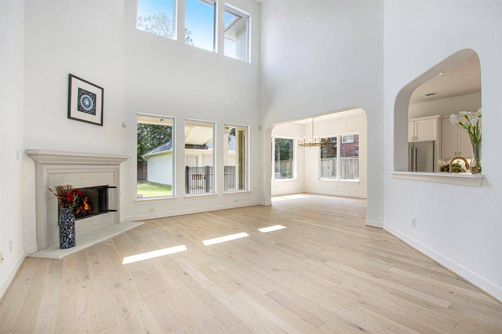 The photo showcases a bright, open living space with high ceilings and large windows. It features a cozy corner fireplace, light wood flooring, and an adjacent breakfast area with views of the backyard.