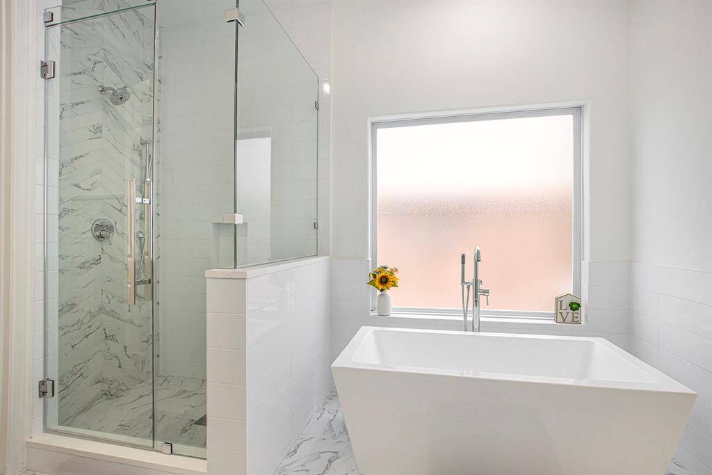 This bathroom features a modern design with a sleek glass-enclosed shower and a freestanding soaking tub. The space is brightened by a large frosted window, complemented by elegant flooring and fixtures.