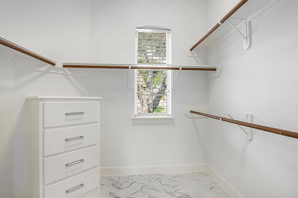 This walk-in closet features ample hanging space with wooden rods, a built-in white dresser with sleek handles, and an awesome  window that allows natural light to illuminate the space. The floor is elegantly finished with stylish tiles.
