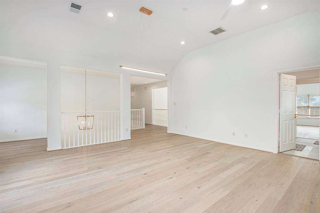 Bright, spacious gameroom with light wood floors, vaulted ceiling, recessed lighting, and a modern chandelier. Features an open layout with white walls and a railing, leading to adjacent rooms.