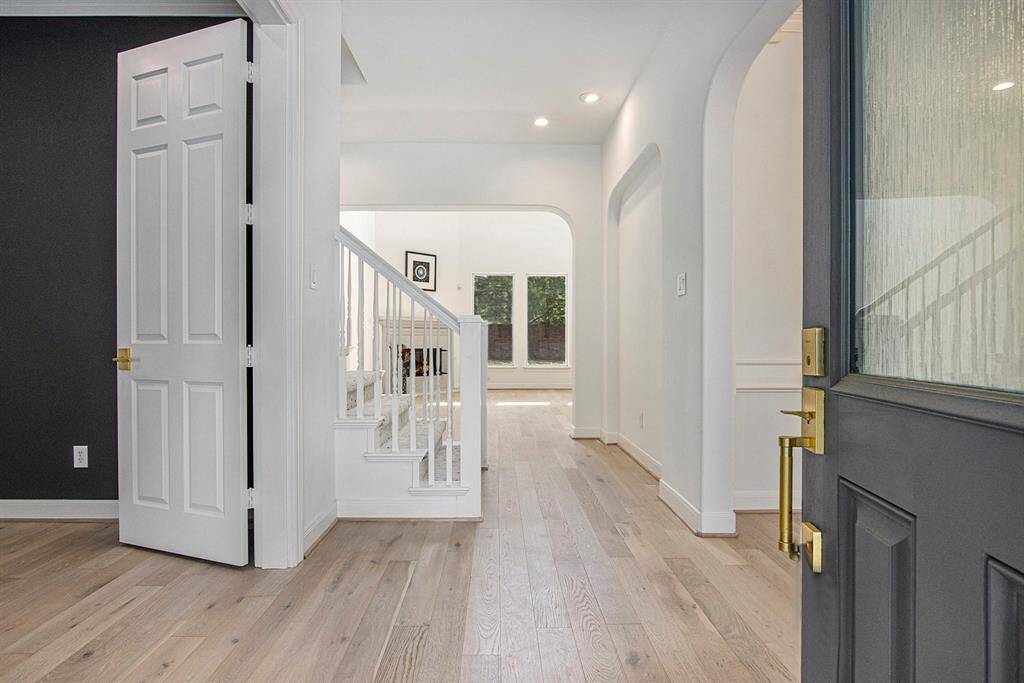Upon entering the home, there is a bright and inviting entryway with white oak wood flooring and modern white walls. The open layout leads to a staircase with white railings and a view of a spacious living room with large windows, allowing natural light to flood in.