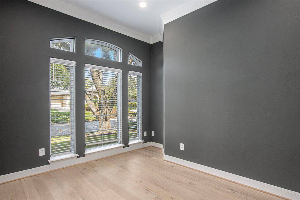 This room is the perfect study room, features large arched windows with blinds, offering ample natural light and a view of the greenery outside. It has stylish gray walls, wood flooring, and white crown molding, creating a modern and elegant space.