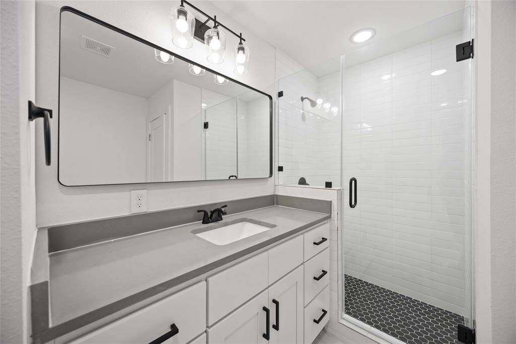 This is a polished bathroom featuring a large vanity with plenty of storage, a wide mirror with overhead lighting, and a walk-in shower with white horizontal tiles and a unique black hexagon-tiled floor.