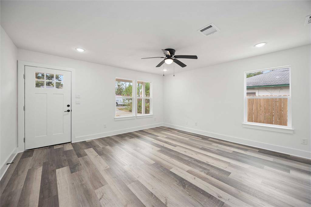 An airy living room with fresh white walls, stylish gray-toned luxury vinyl flooring, ample natural light from large windows, and a ceiling fan for comfort.