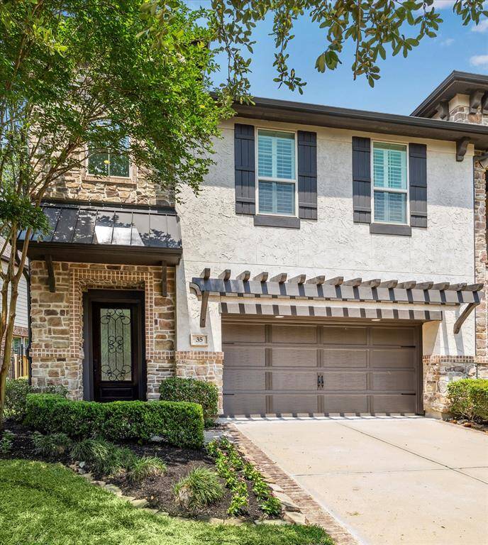 Elegant two-story residence showcasing a sophisticated blend of stone and textured stucco, accented by rich wood shutters and a sleek pergola above the carriage-style garage. A wrought-iron front door and manicured landscaping complete the upscale curb appeal.