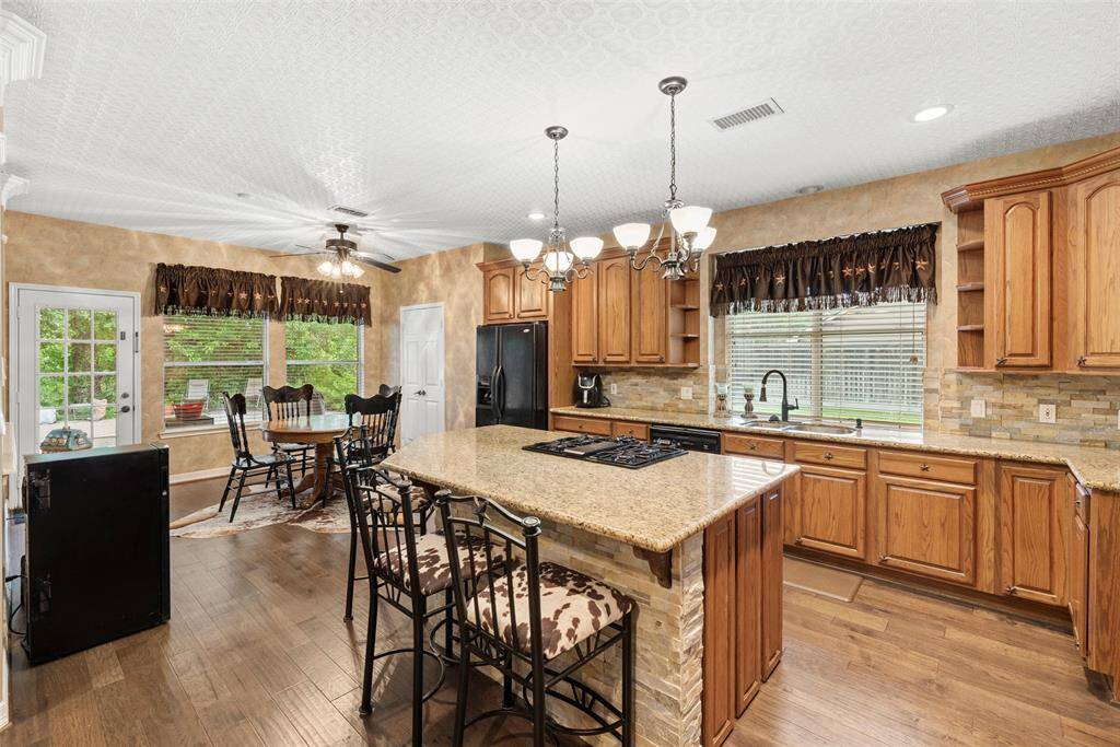 Open kitchen features warm wood cabinets, a large central island with a cooktop, and elegant lighting fixtures. It opens to a cozy breakfast area with large windows and backyard access, providing a bright and inviting atmosphere.