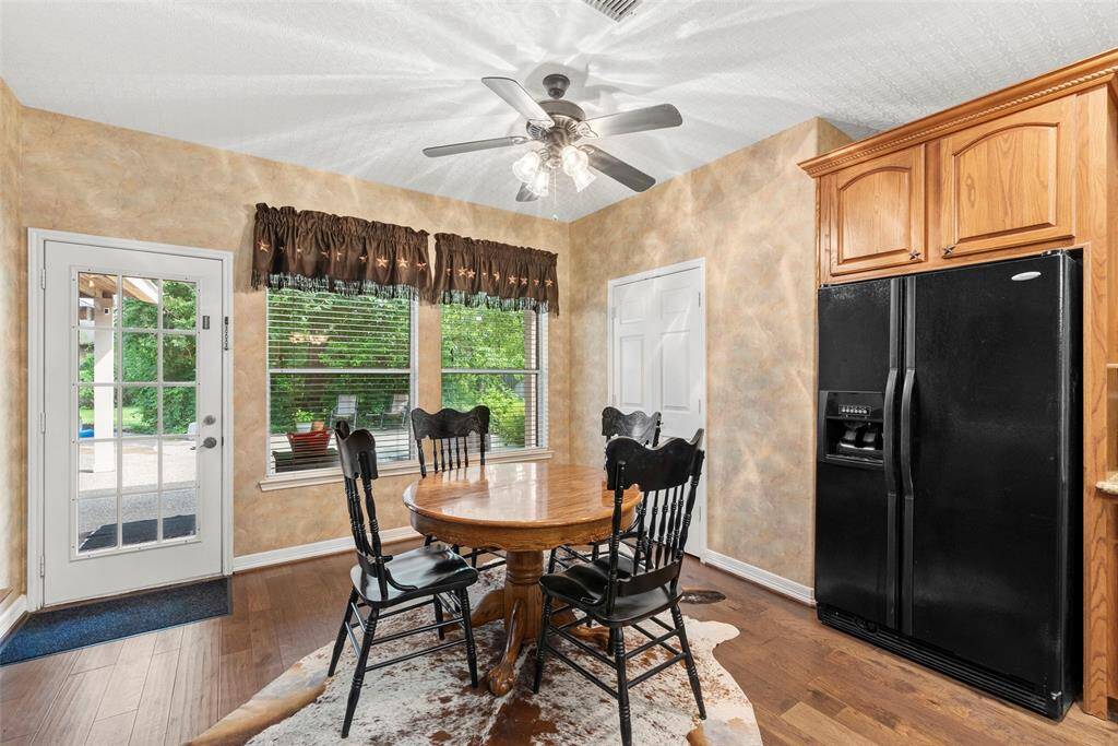 A cozy dining area that features warm, textured walls, a ceiling fan, and ample natural light from a large window. A black refrigerator and wooden cabinets and pantry offer practical kitchen storage. The space opens to the outdoors through a glass door, allowing for more natural light.