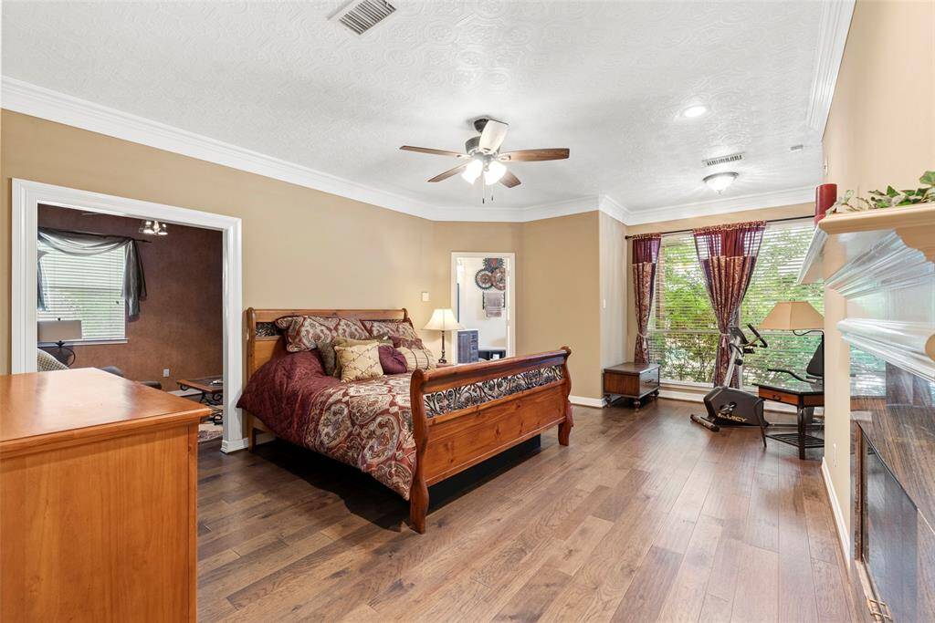 This spacious primary bedroom features hardwood floors, a ceiling fan, and ample natural light from large windows. It includes a cozy seating area and has a warm, welcoming ambiance with neutral wall colors and elegant crown molding.