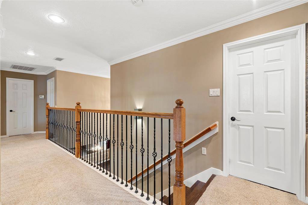 This photo shows a bright upstairs hallway leading to game room and secondary bedrooms with a carpeted floor, featuring two white doors and a staircase with a wooden railing and decorative iron spindles. The walls are painted a warm beige, and there's recessed lighting in the ceiling.