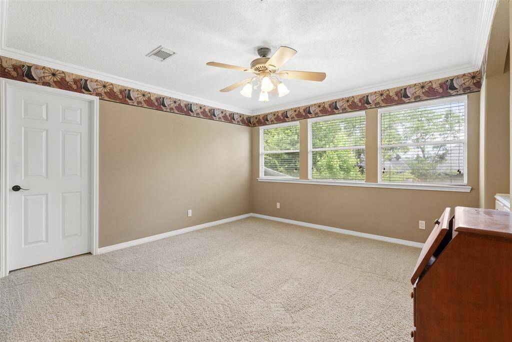 Bright and spacious game room with neutral walls, carpet flooring, and a ceiling fan. Large windows provide natural light and a view of greenery.