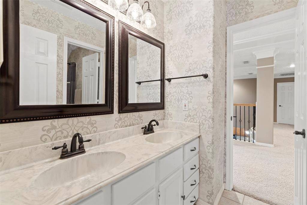 This bathroom features a double vanity with elegant marble countertops and dark framed mirrors. It has stylish wallpaper and modern fixtures, offering a luxurious feel. The space opens to a carpeted hallway with a view of a staircase and to secondary bedroom.