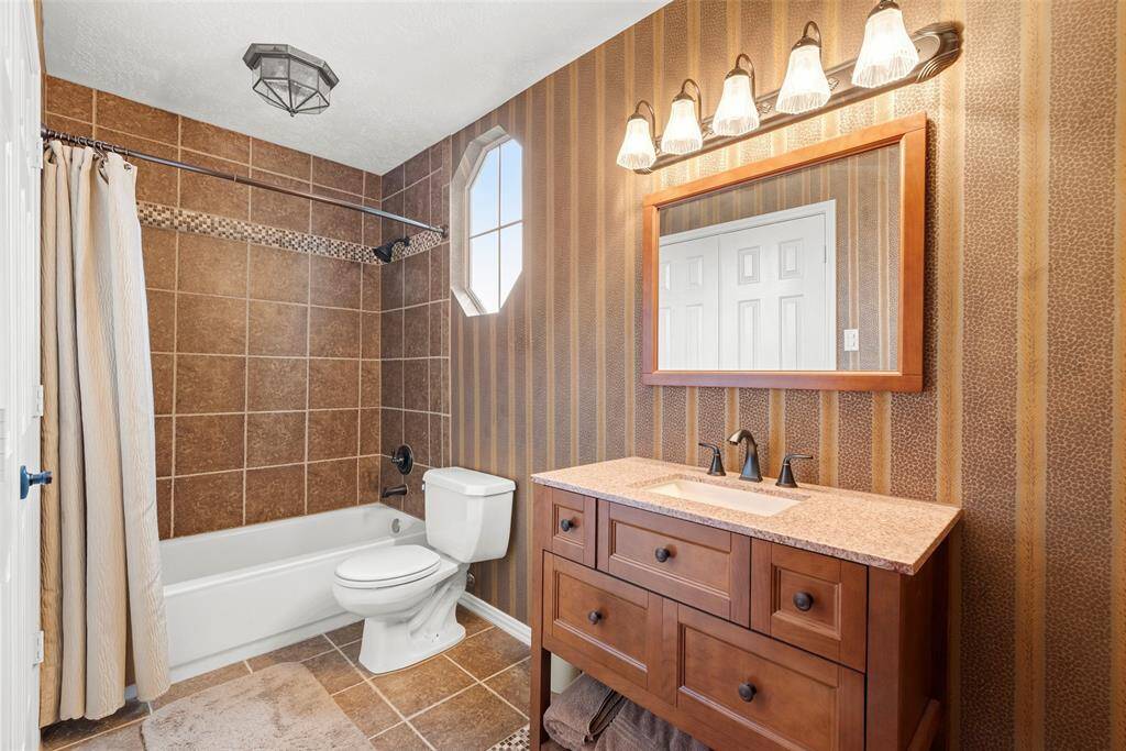 Third bathroom features warm, earth-toned tiles and walls, a classic white bathtub with a shower and a wooden vanity with a granite countertop. The space is well-lit with stylish light fixtures above a large mirror.