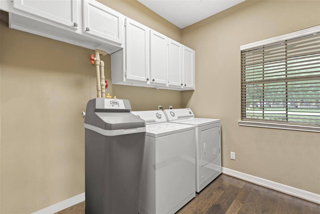 This photo shows a well-lit laundry room with a washer, dryer, and water softener which are all included with house.  It features white cabinetry for storage and a window with blinds providing natural light. The space has neutral walls and wood flooring.
