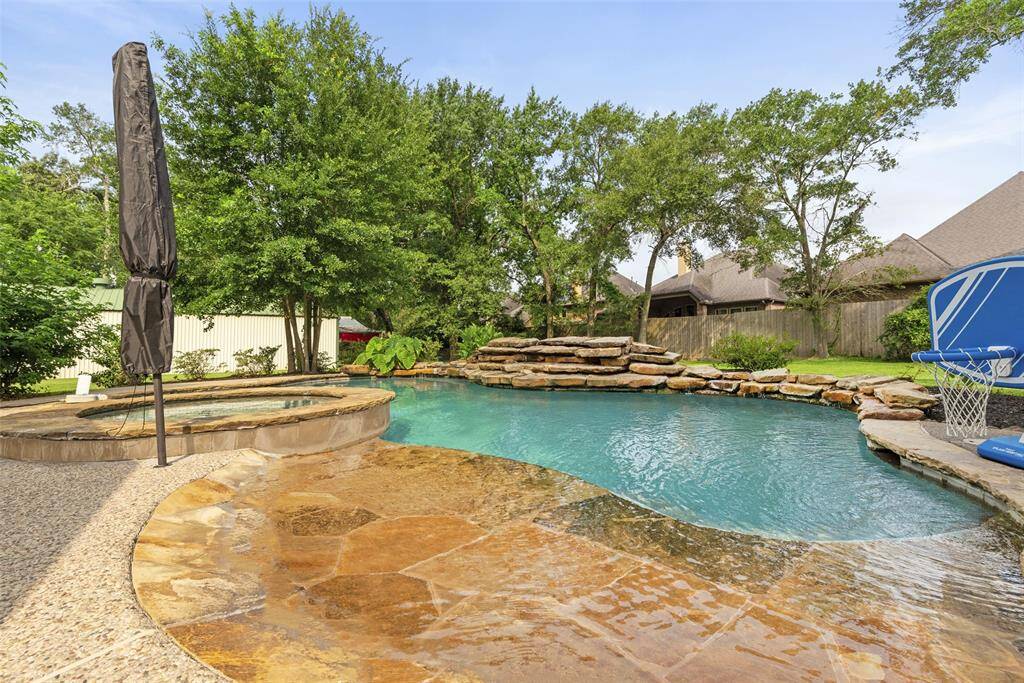 This inviting backyard features a beautiful pool with a stone waterfall, surrounded by lush greenery and trees for privacy. A spa area and a basketball hoop add to the recreational appeal, making it perfect for relaxation and entertainment.