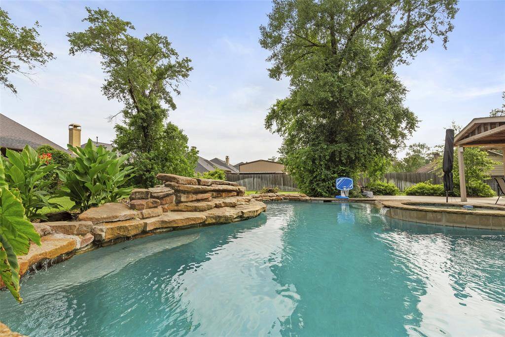 Inviting pool with natural stone accents and lush greenery, creating a private oasis. There's a covered patio area ideal for relaxing or entertaining.