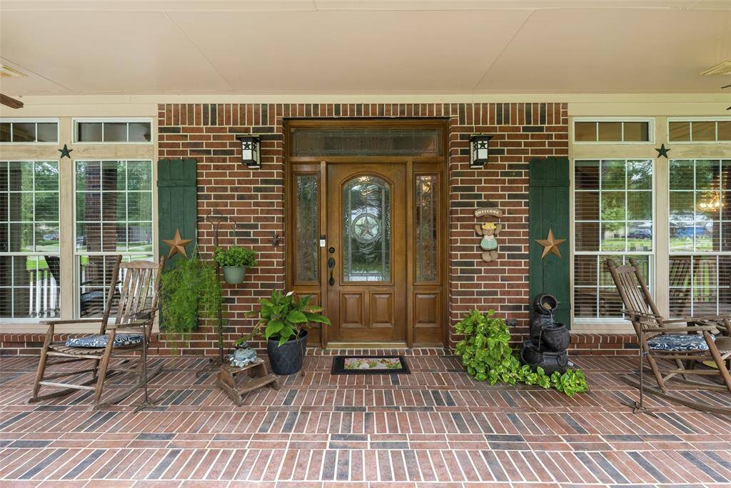 Welcoming front porch with a classic brick exterior, featuring a wooden door with decorative glass panels. Large windows with white trim enhance the natural light and provide a view of the surrounding greenery.