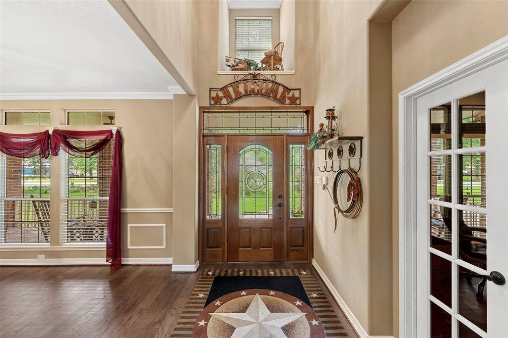This inviting entryway features a grand wooden double door with decorative glass panels, high ceilings, and abundant natural light. Elegant crown molding and tasteful decor add charm, while a glass-paned door to the right leads to study/office.  The space is warm and welcoming, perfect for making a memorable first impression.