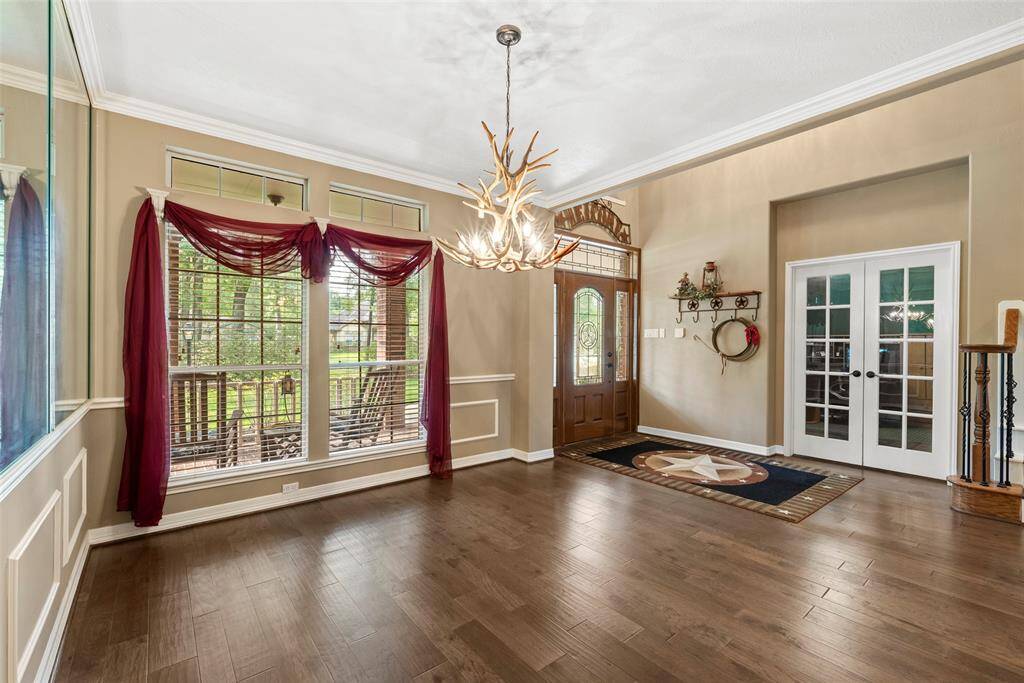 This spacious entryway features elegant hardwood floors, large windows providing ample natural light, and a striking antler chandelier. The area includes a decorative front door, mirrored walls, and French doors leading to study/office, creating a welcoming and stylish atmosphere.