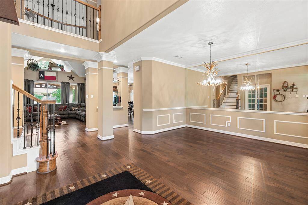 Home features an open floor plan with elegant wood flooring, high ceilings, and decorative crown molding. The large living area flows into a dining space with modern chandeliers and mirrored wall accents, creating a bright and inviting atmosphere. A stylish staircase adds character, leading to an upper level.