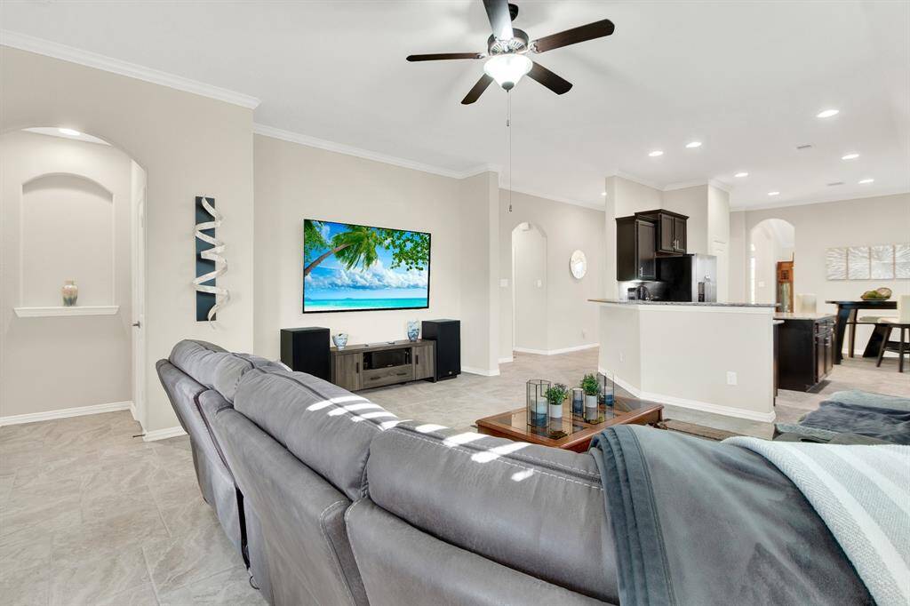 Spacious open-concept living area with modern tile flooring, neutral walls, and recessed lighting. Features a cozy sectional, large TV, ceiling fan, and a sleek kitchen with dark cabinetry. Ideal for comfortable living and entertaining.