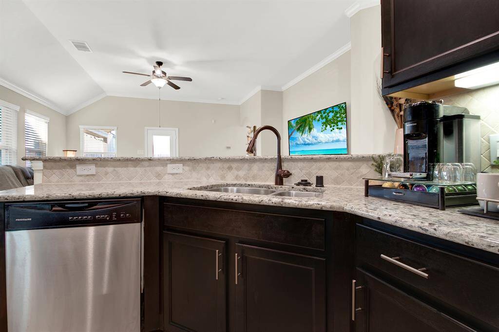 Modern kitchen with granite countertops, stainless steel appliances, and dark wood cabinets. It features an open layout with a view into the living area, which has large windows and a ceiling fan.